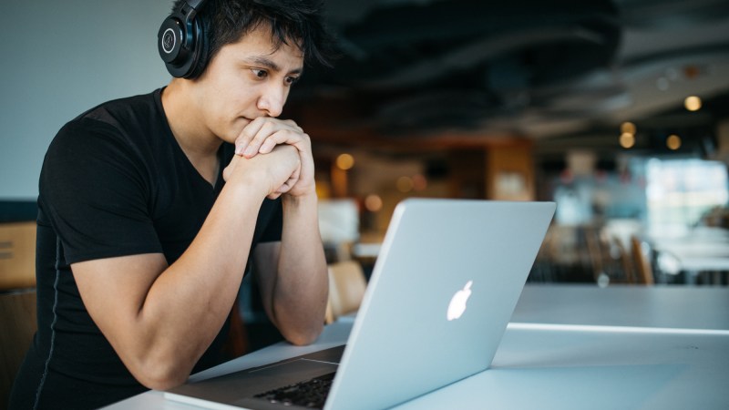 A man wearing black headphones and a black t-shirt sitting in front of a silver Macbook laptop with his elbows on the table and his hands clasped in front of his mouth, looking concerned.