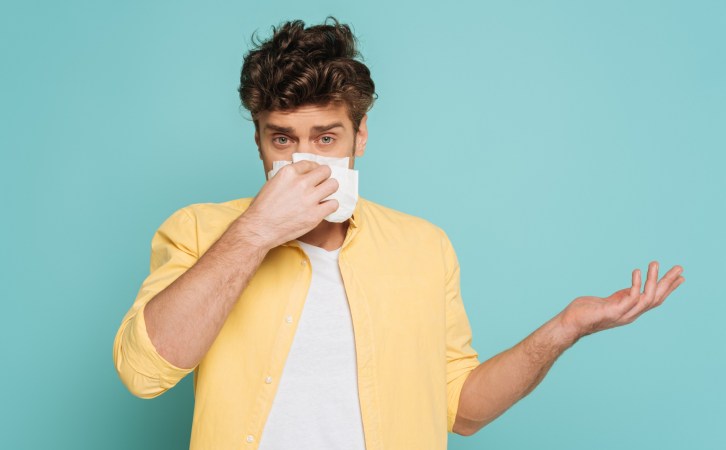 Person with short brown hair and bright yellow shirt on a light blue background holding tissue up to runny nose