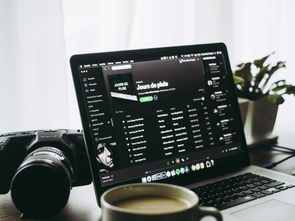 A laptop on a desk with Spotify on the screen. A camera, a plant, and a full mug of coffee share the desk with the laptop.