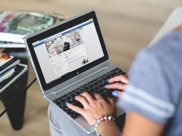 A person using Facebook on a laptop while sitting in front of a table with books on it.