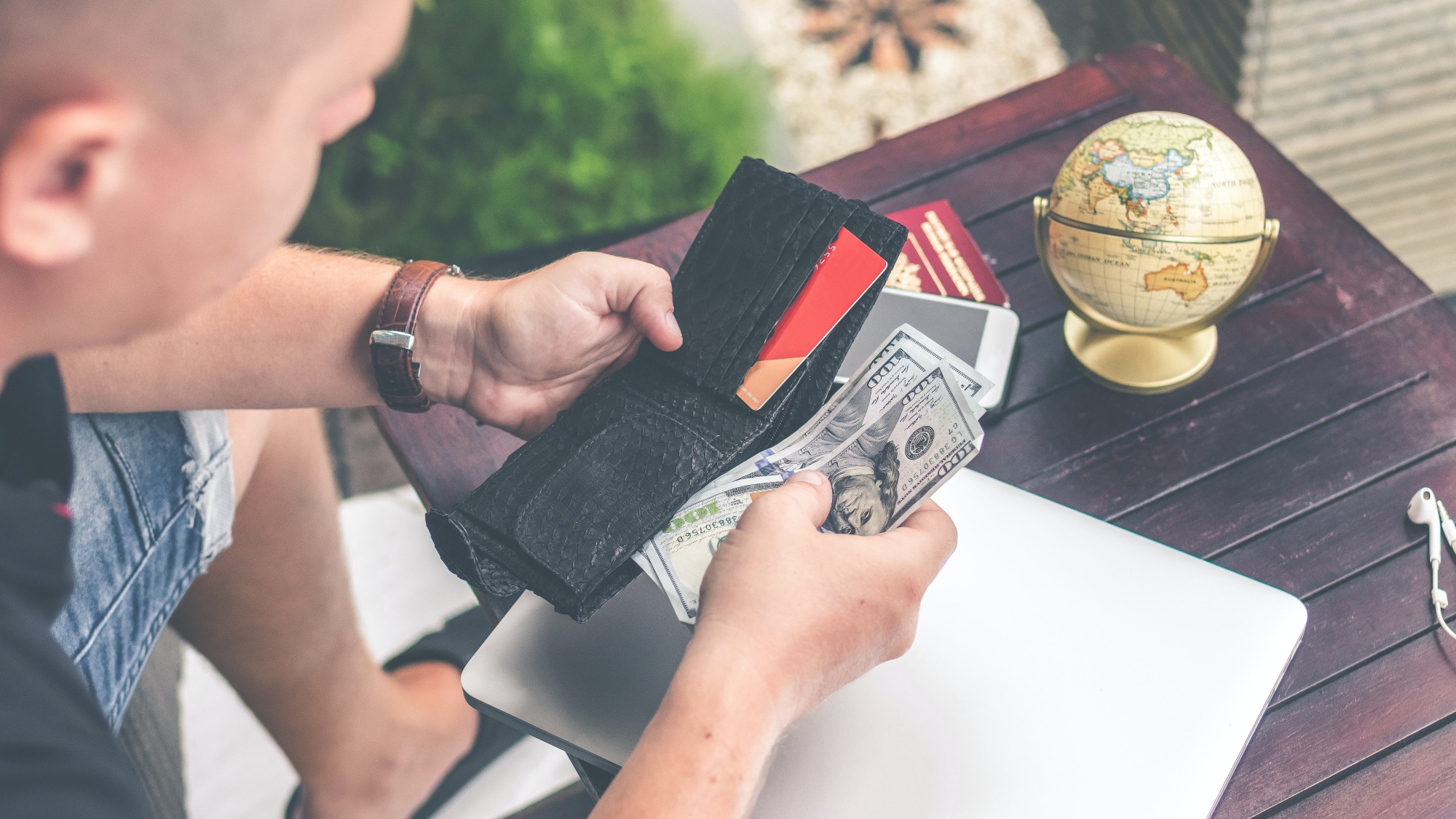 A man sitting and opening his wallet to remove several hundred dollars in cash with a laptop on the table in front of him.