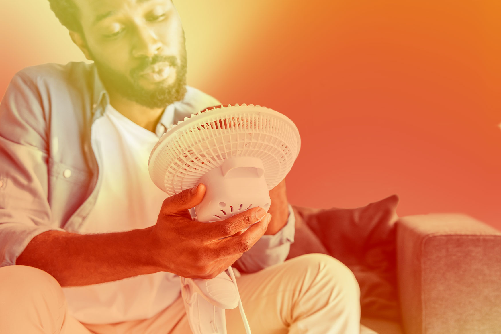 A man sitting and holding an electric fan in front of his face.