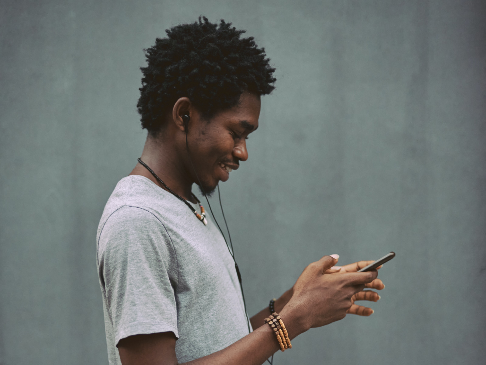 A person using finger gestures on their phone while wearing headphones and standing in front of a gray wall.