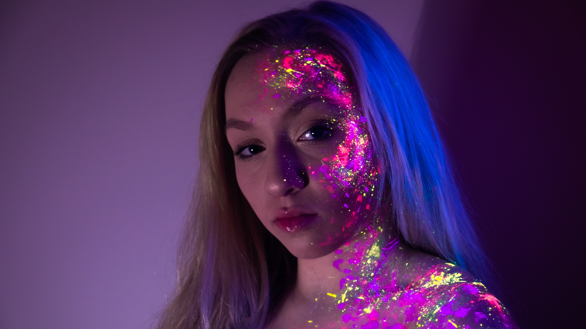 A woman wearing ultraviolet makeup or body paint on half her face, standing under a blacklight.