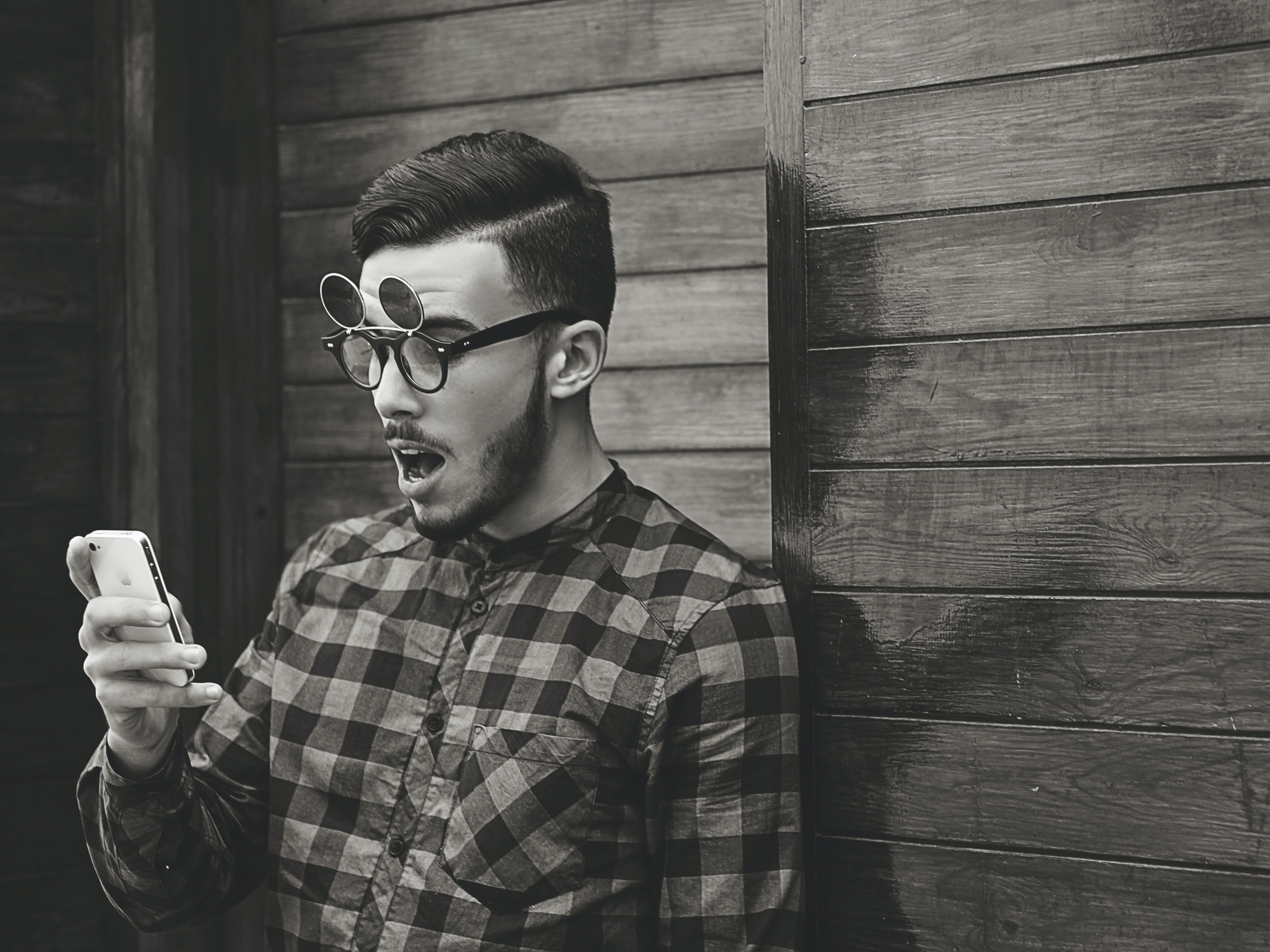 A young man in a plaid shirt standing against a wooden wall wearing glasses with flip-up outdoor lenses, looking at his phone with a shocked face.