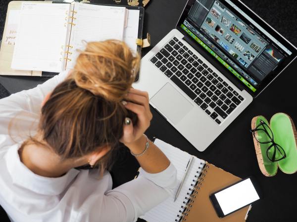 A busy woman overwhelmed at work, holding her face in her hands in front of a Macbook laptop with some open notebooks around it.