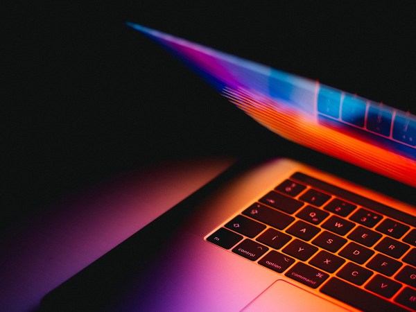 A partially closed Macbook laptop emitting multi-colored light in a dark room.