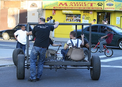 A group of men pedaling and pushing a pedal-powered replica tank frame down a street.