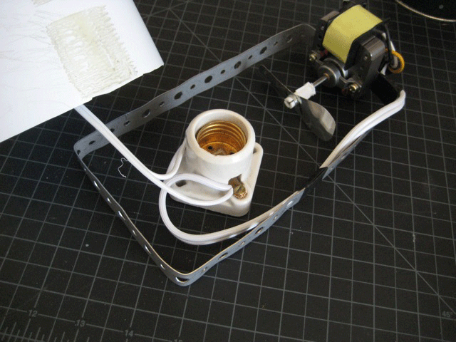 A light bulb socket and a motor with a propeller inside a bracket on top of black grid paper.