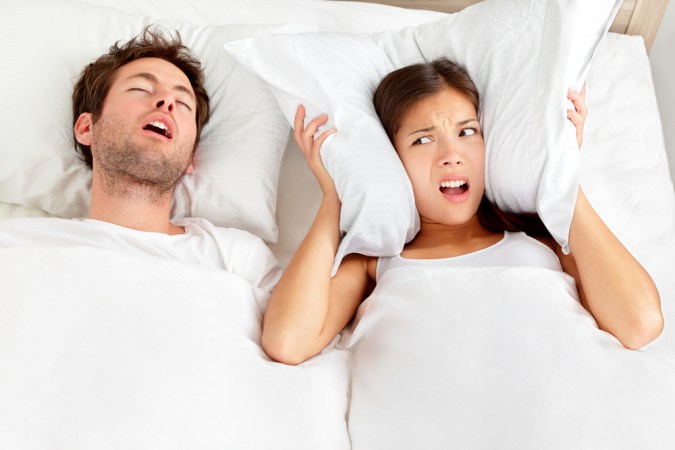 A man snoring in a white bed, while a woman angrily holds a pillow over her ears next to him.