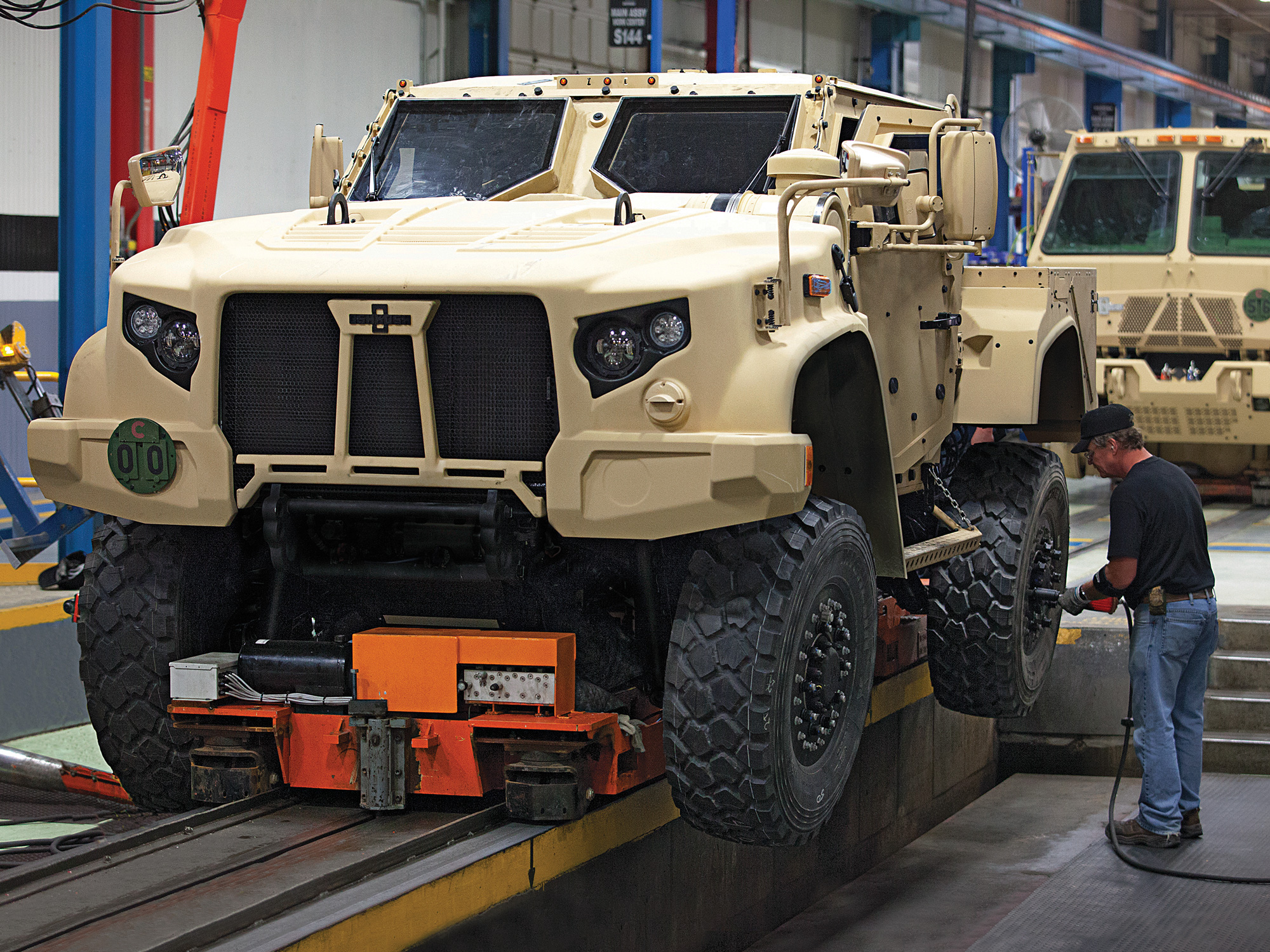 A beige heavy-duty armored car in a shop with a white man in jeans working on it