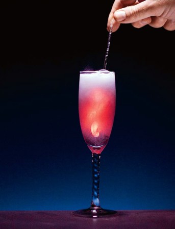 A person using a metal cocktail stirrer to mix chlorine and hydrogen peroxide in a champagne glass. The liquid is foaming and glowing red-orange.