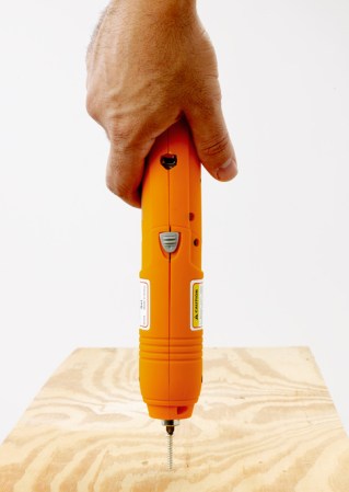 A hand holding an orange cordless screwdriver that's on top of a screw partially sunken into a sheet of plywood.