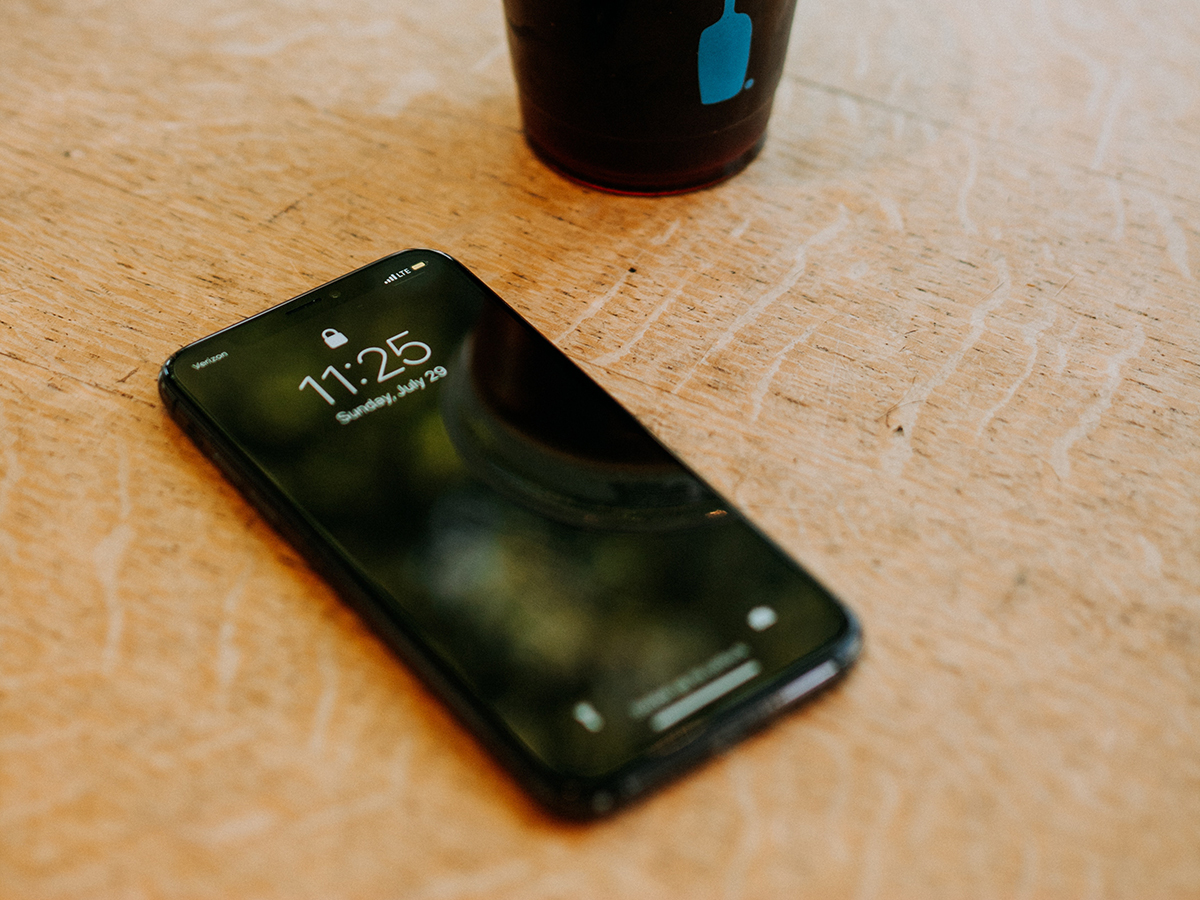 An iPhone on a wooden table at a coffee shop with a coffee cup next to it.