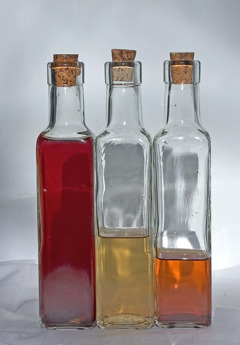 Three thin rectangular cork-plugged bottles in a row, the left one nearly full of red vinegar, the middle one half full of yellow vinegar, and the right one about a quarter full of orange vinegar.