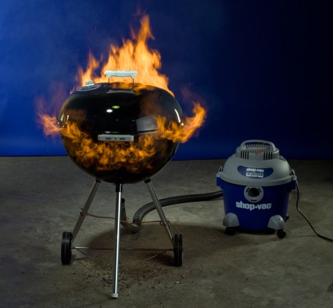 A black charcoal grill with its lid on, billowing flames. There's a vacuum cleaner attached to its underside.