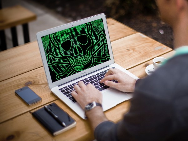 A person sitting in front of a laptop that has a skull and crossbones in green code on the screen, indicating that it may have been infected with malware that they'll now need to remove.