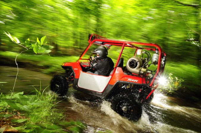 A man wearing a black firesuit and black helmet driving a jet turbine-powered ATV through a stream in the woods.