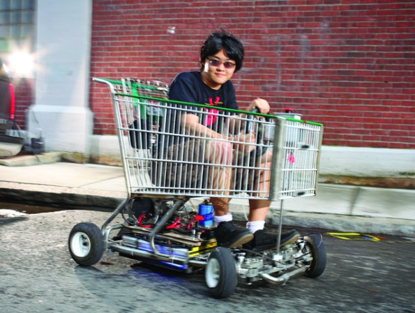 A college student wearing sunglasses and a black t-shirt sitting in a go-kart that's built out of a shopping cart.