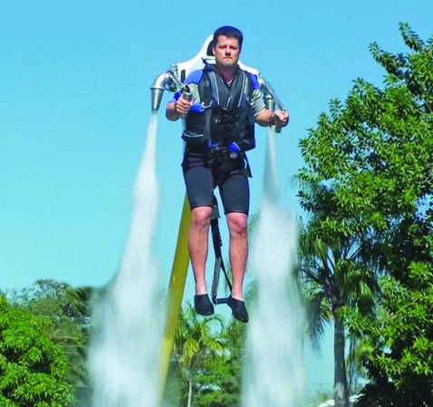 A man using a water-powered jetpack to rise into the air near some trees under a blue sky.