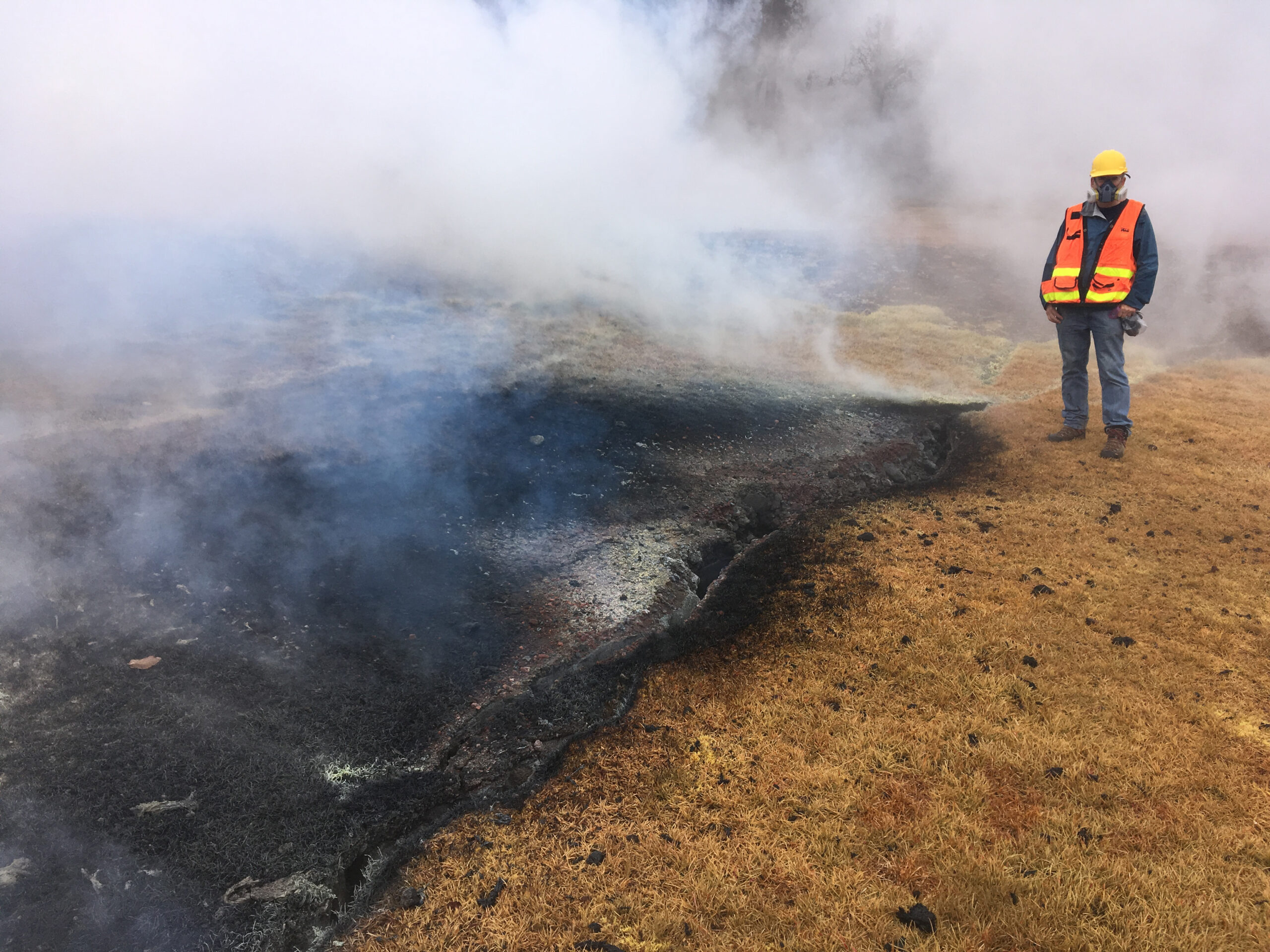 Man in vest and mask standing next to burning ground.