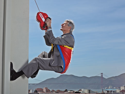 An old man in a gray suit using a rappelling device to descend from a tall office building.