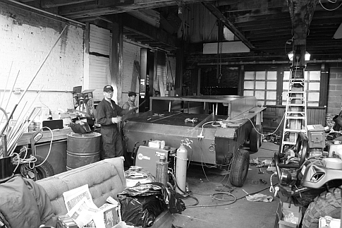 A pedal-powered replica tank frame in a garage, partially covered with cladding. Grayscale.