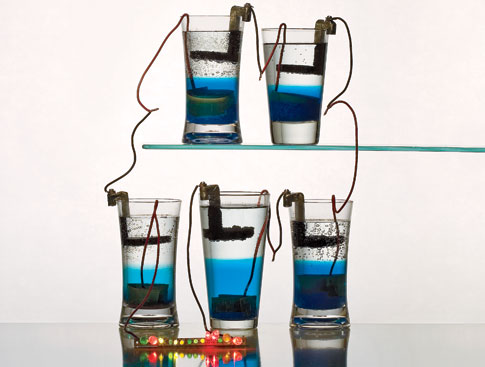 Five drinking glasses filled with clear liquid on top and blue liquid on the bottom, linked together to create a gravity-cell battery.