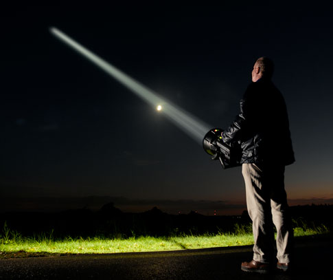 Ralf Ottow stands outside at night, pointing a huge beam of light into the sky, with the moon in the background.