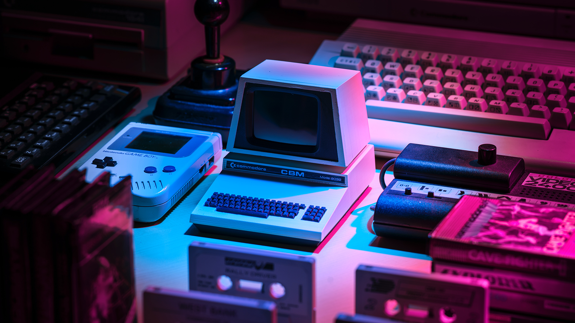 Vintage gadgets like computers, keyboards, and a GameBoy sit on a table under pink and blue lights.