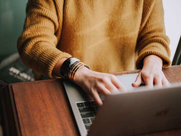 A person in a tan sweater sitting at a laptop computer on a wooden table.