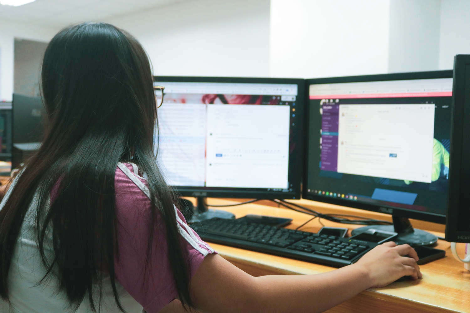 A black-haired woman with glasses using a computer with two monitors—an excellent use-case for Windows keyboard shortcuts.