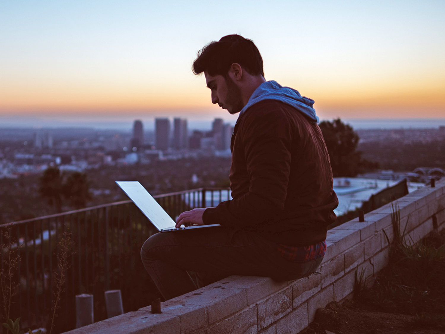 A man working on a laptop while sitting on a rock overlooking a city at sunset.