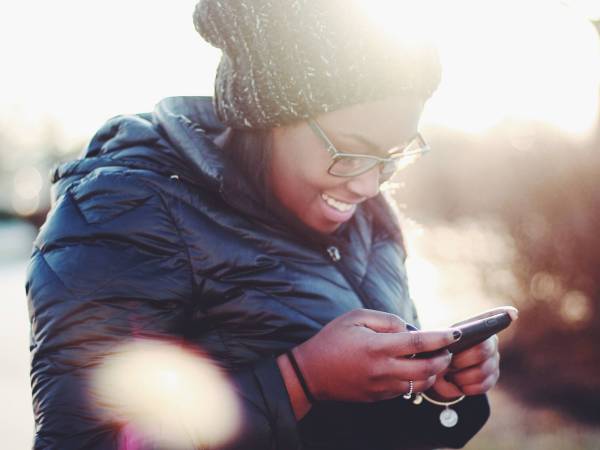 A person standing outside wearing a blue winter coat and a gray knit hat while smiling and looking at their phone.