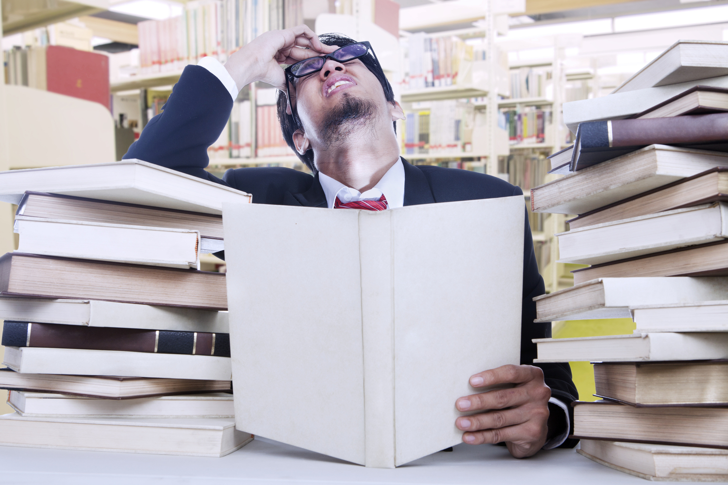 a stressed man in a suit and glasses trying to read a lot of books in a library