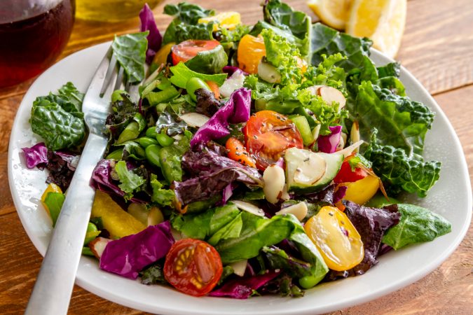 Close up of fresh organic super food salad in white bowl with fork on side and olive oil, red wine vinegar and lemons