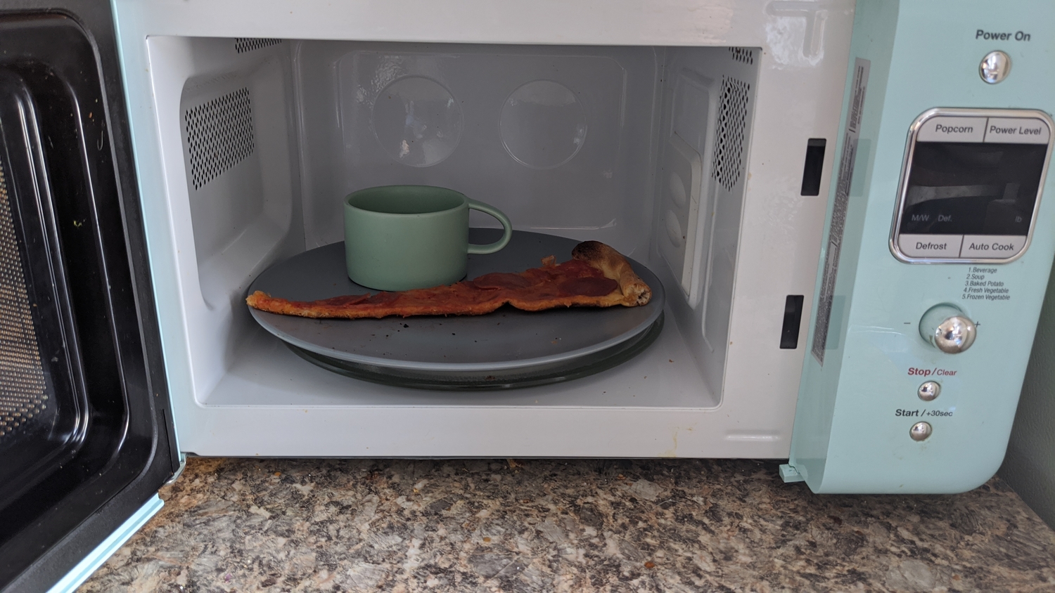 A slice of cheese pizza in a teal microwave with a mug of water.