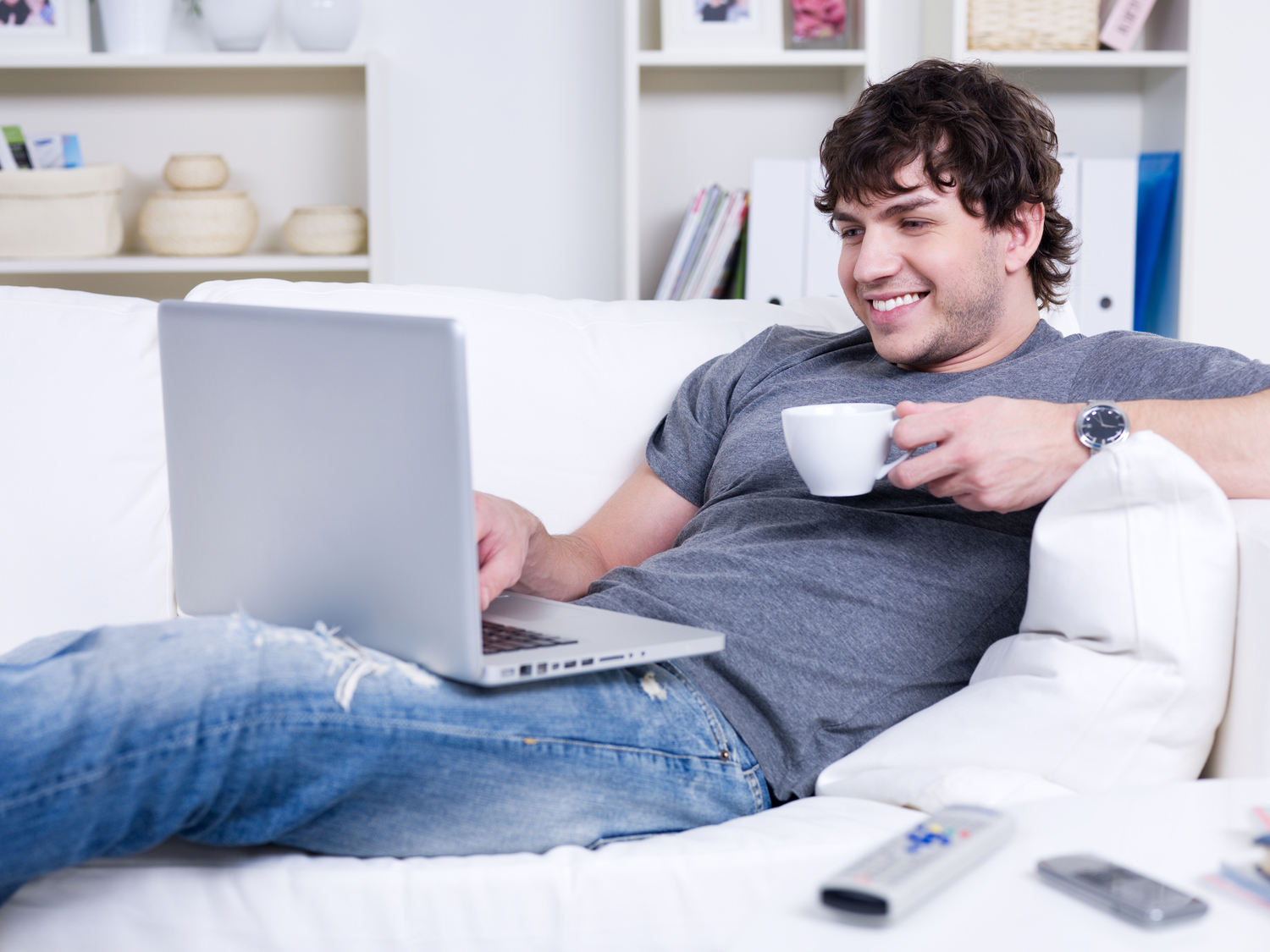 a man on his white couch with a cup of tea or coffee, smiling while using his laptop