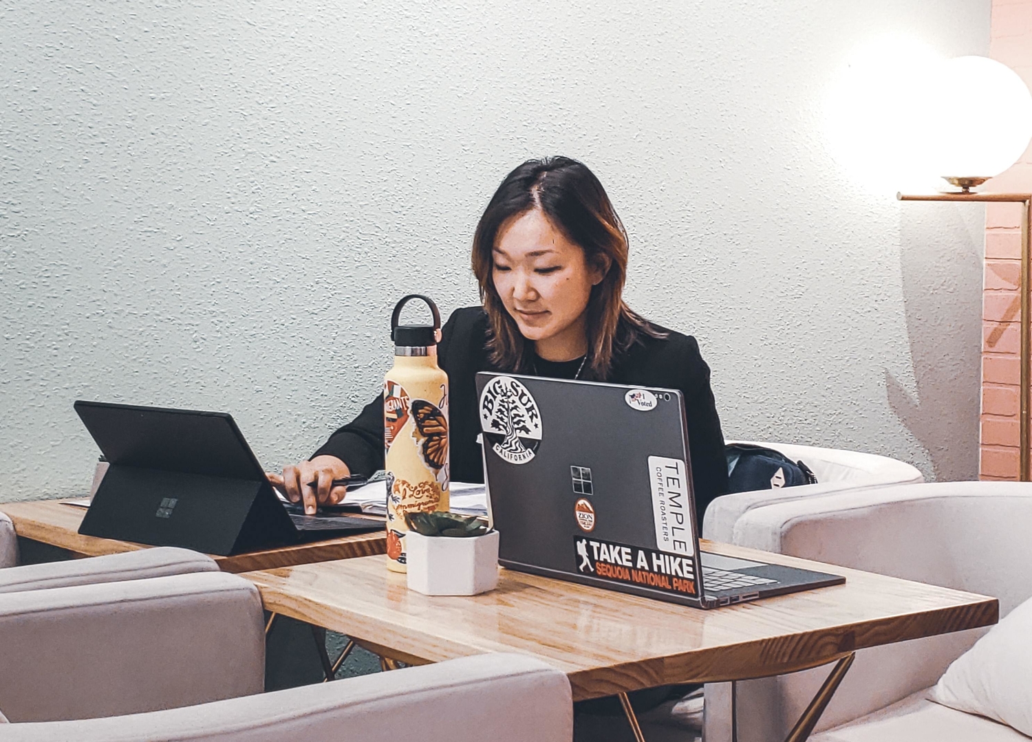 a woman at a wooden table using a Windows tablet or laptop next to another Windows laptop