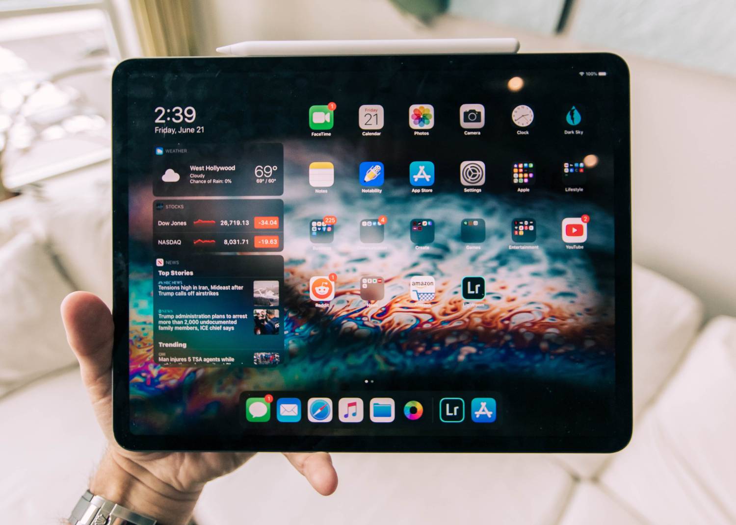 A hand holding an iPad over a white bed in a sunlit bedroom.