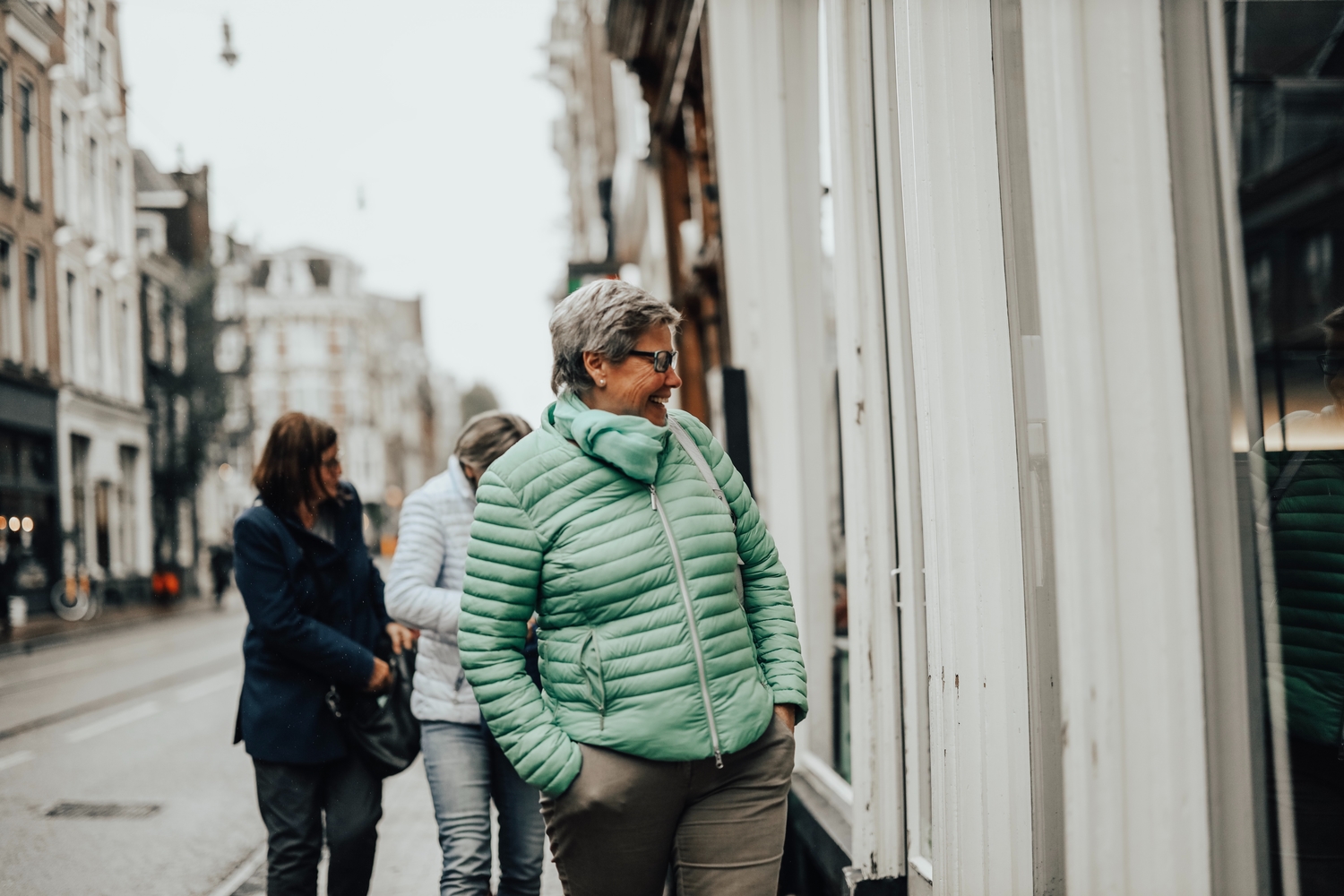 a woman in a green winter jacket walking on the street and looking in a window while smiling