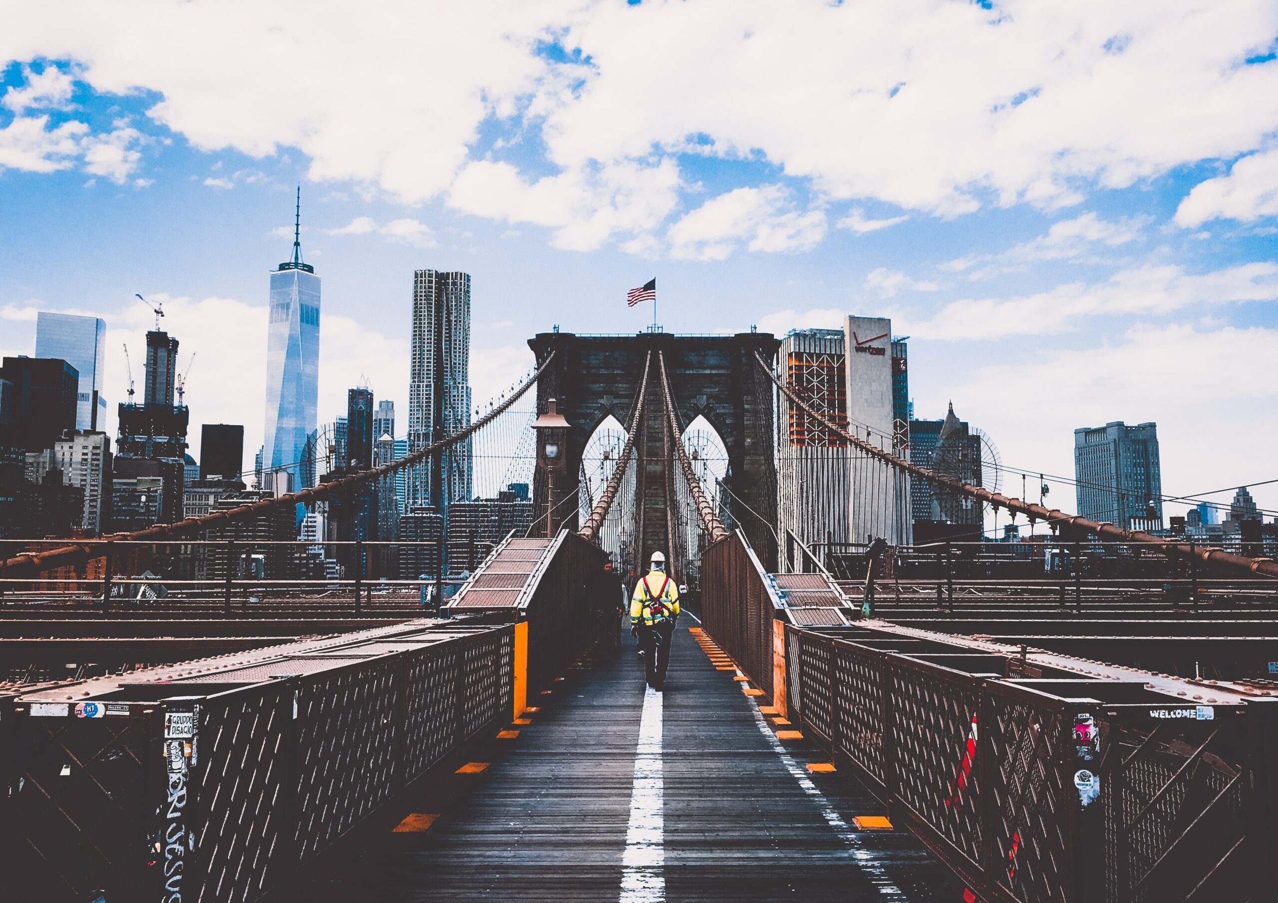 a man walking alone on the brooklyn bridge with the NYC skyline and an American flag visible