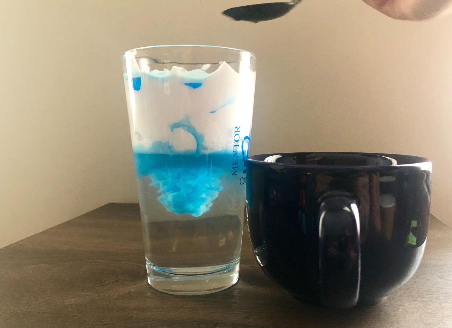 a photo of a person pouring colored water into a glass of shaving cream and water to demonstrate rainfall with a science experiment project creating a storm in a glass