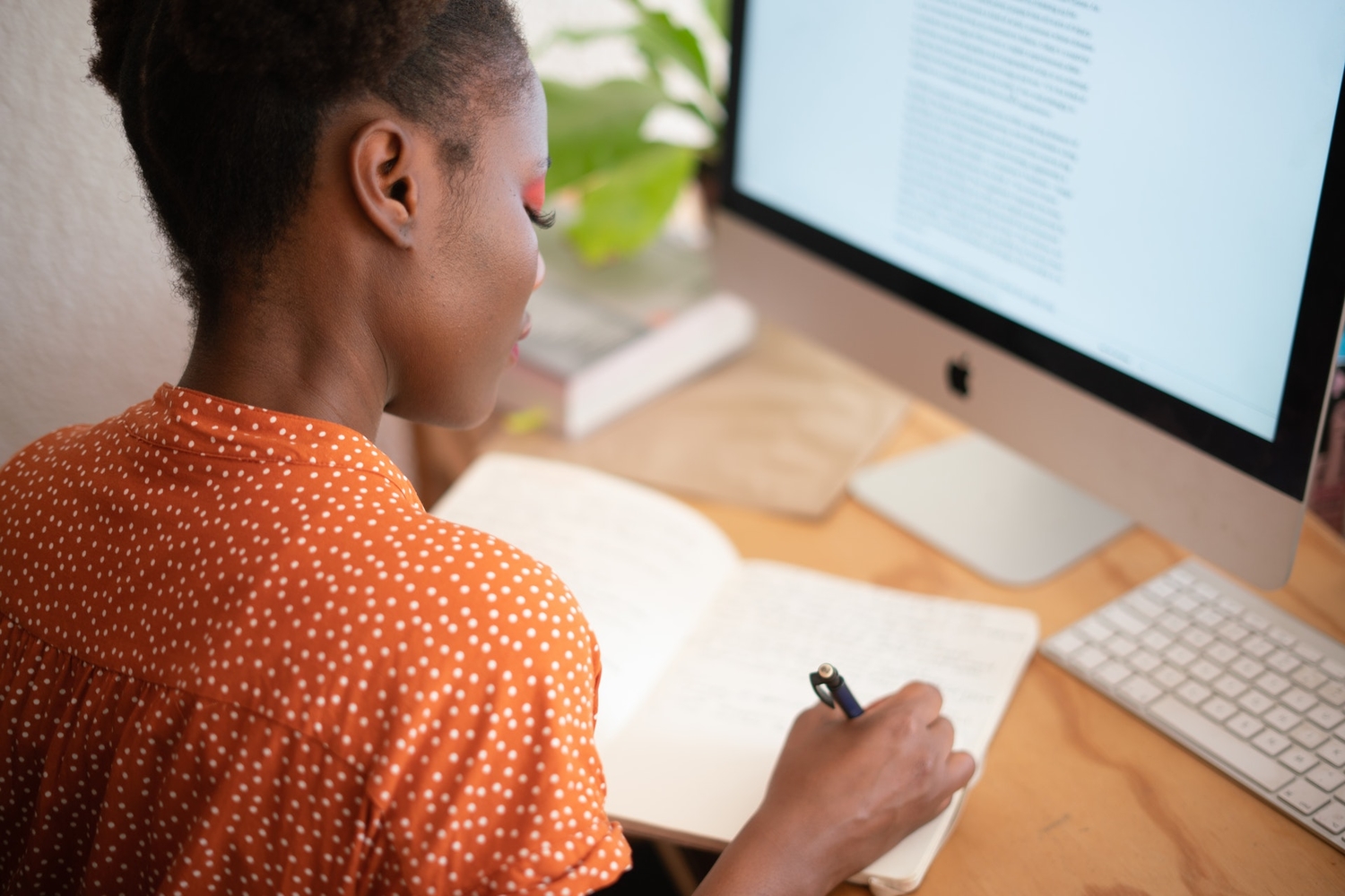 a woman writing notes in a book in front of a computer while working from home