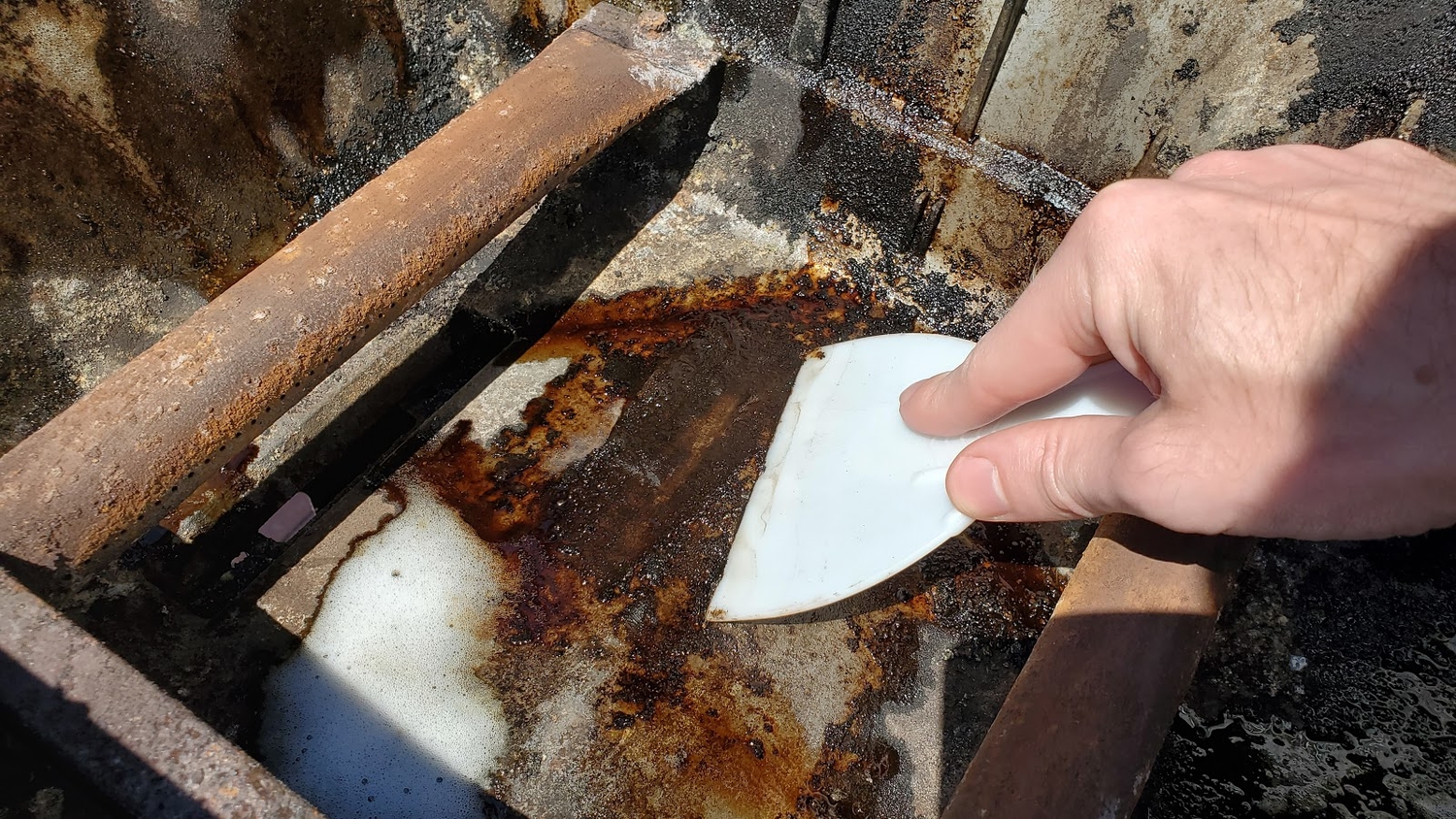 a person scraping debris out of a grill cookbox with a plastic paint scraper and some grill cleaner