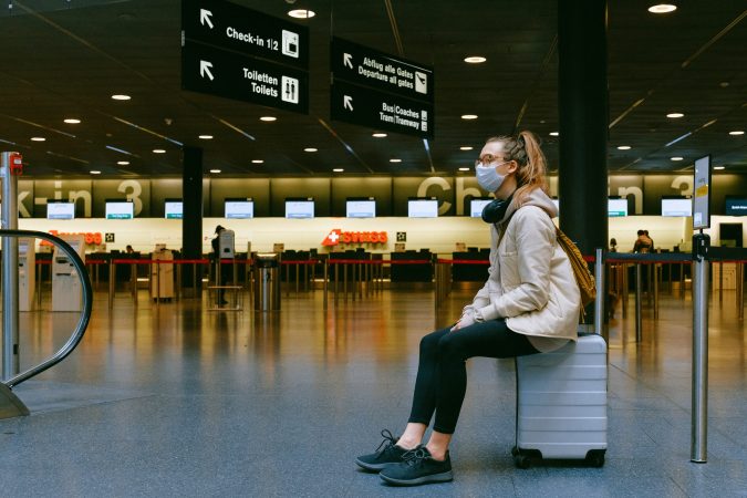 woman wearing mask sitting on suitcase in airport