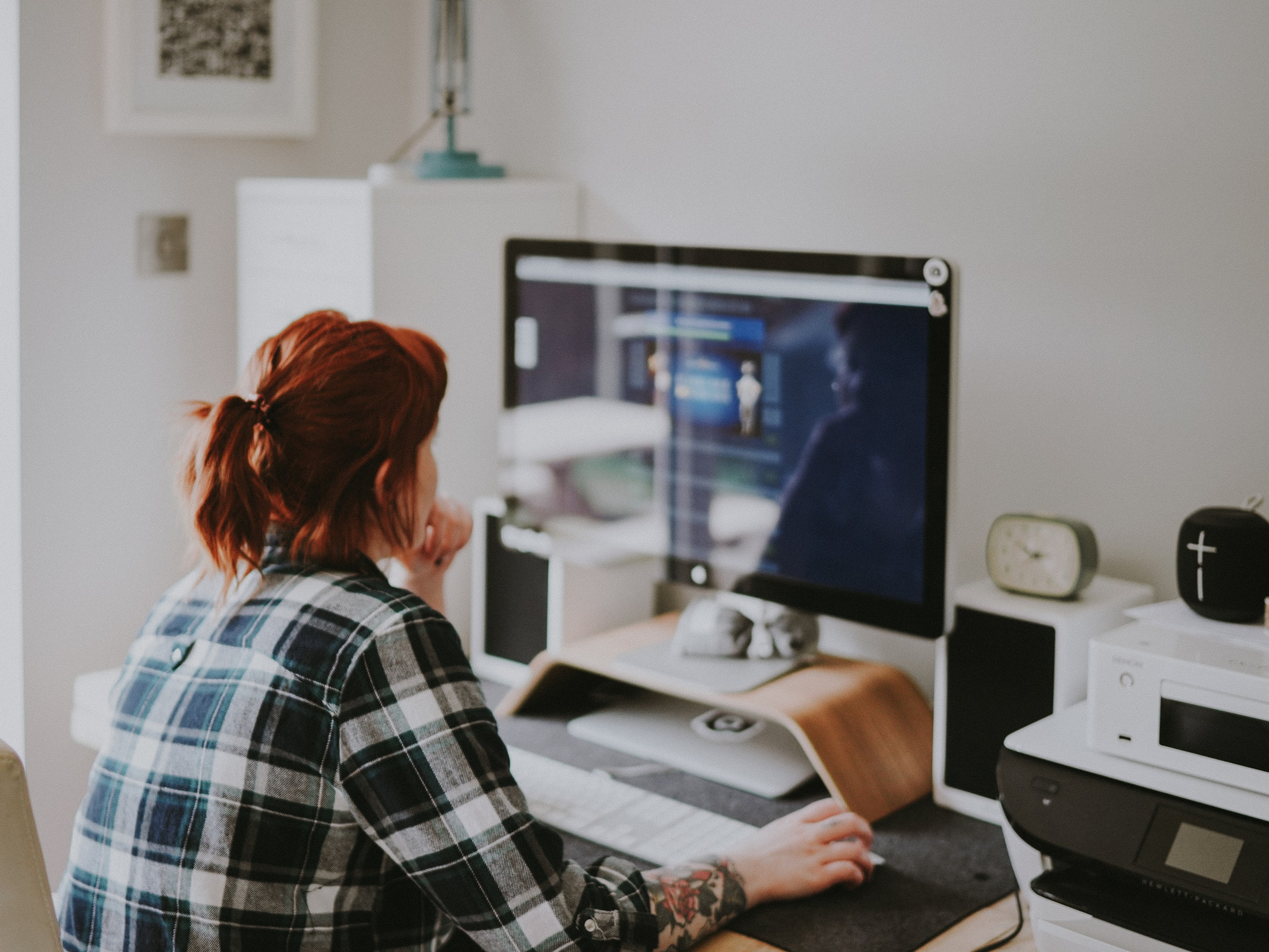 A redheaded person in a blue plaid shirt sitting at a desktop computer, doing work and trying to minimize distractions while working from home.