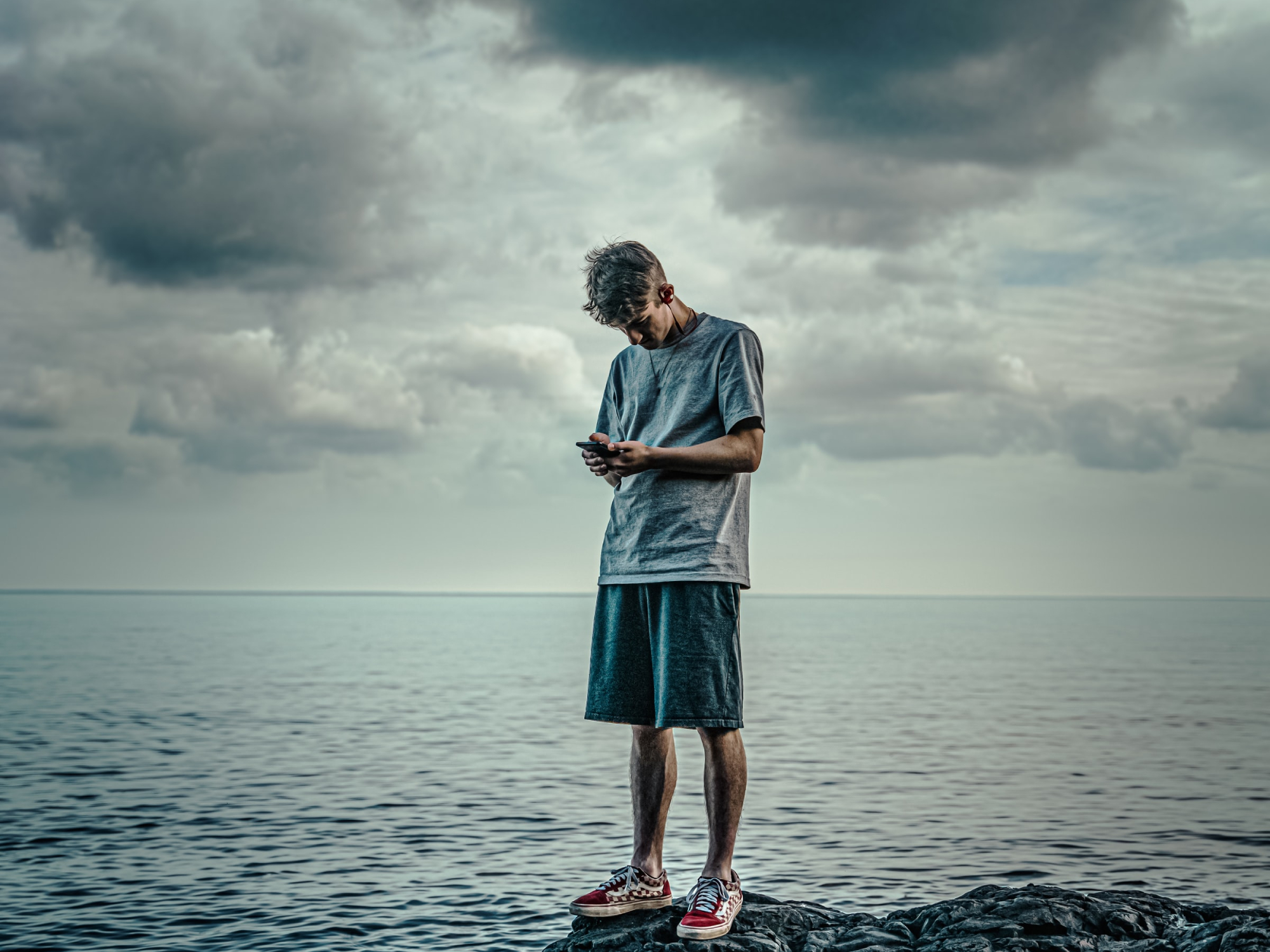 A person standing on a rock by the water under cloudy skies while looking at his phone—hopefully he has a good weather app.