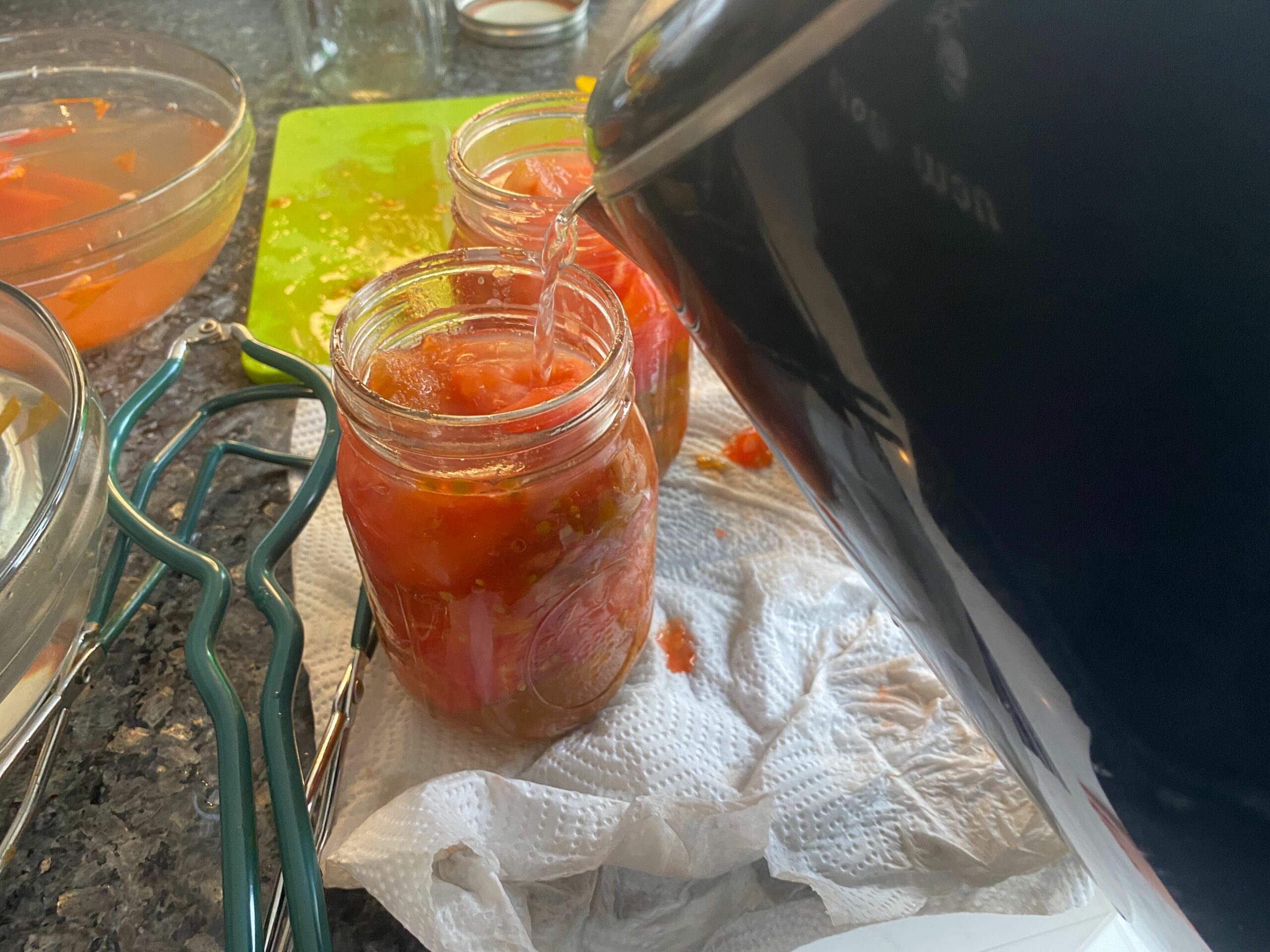 a person pouring boiling water from a kettle into a jar of sliced tomatoes