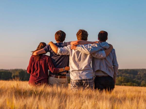 Four people standing facing away from the camera, with their arms around each others' shoulders, in the middle of a field of golden grass under a blue sky.